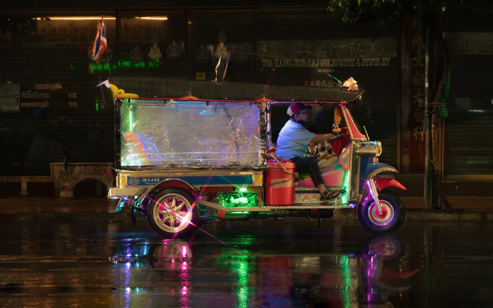 A tuk-tuk driving through a rainy street with city lights on the wet road